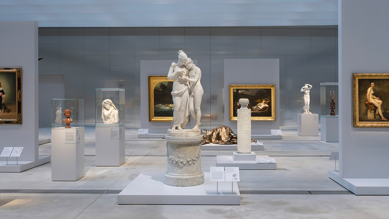 Vue d'une section de la Galerie du Temps au Louvre-Lens, mettant en avant une sculpture en marbre représentant deux figures féminines enlacées, entourée de bustes, statues et peintures encadrées, exposées dans un espace lumineux et épuré.
