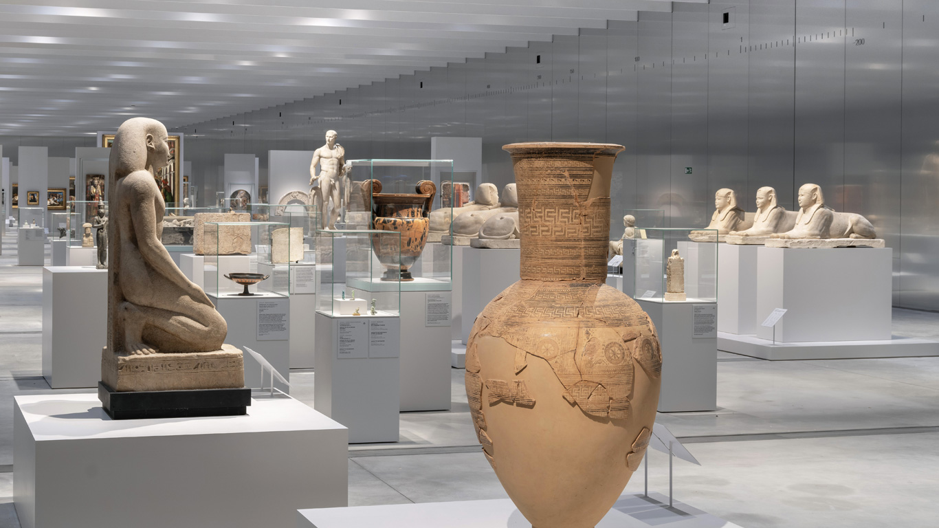 Vue de la Galerie du Temps au Louvre-Lens, présentant une grande amphore ornée de motifs géométriques et de gravures, une statue égyptienne en position agenouillée, ainsi que des sphinx et diverses œuvres antiques exposées dans des vitrines sous un éclairage moderne.