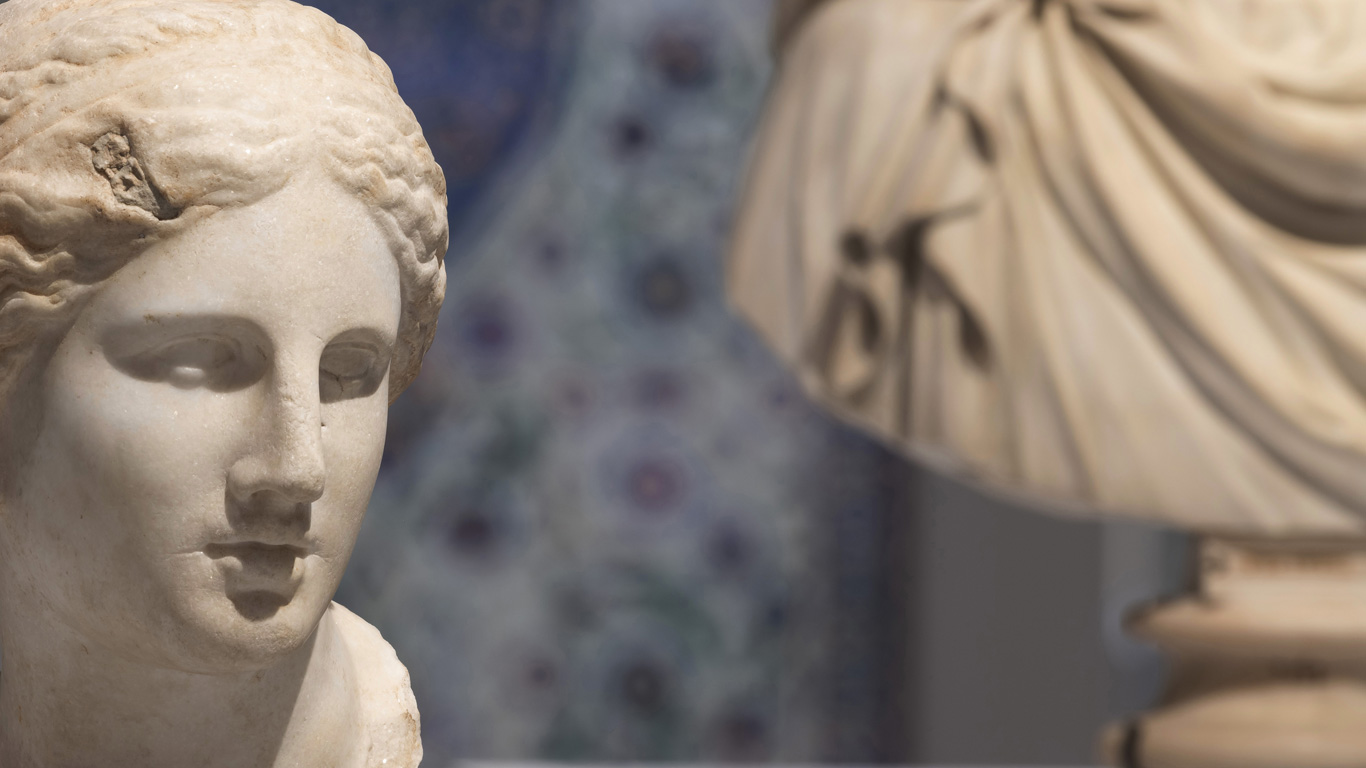 Gros plan sur une sculpture antique en marbre représentant une tête de femme aux traits doux et aux cheveux ondulés, avec en arrière-plan une draperie sculptée et un décor ornemental flou, exposée dans la Galerie du Temps au Louvre-Lens.